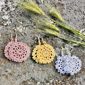 May include: Three pairs of crocheted earrings in pink, yellow, and white. The earrings are round and have a delicate, intricate design. They are hanging from silver hooks.