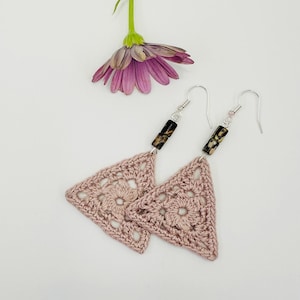 May include: A pair of handmade, light pink crochet triangle earrings with a floral design. The earrings have silver-colored hooks and are accented with dark brown and tan wooden beads. A purple daisy with a green stem is in the background.