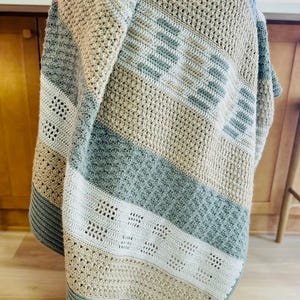 May include: A crocheted throw blanket with a striped pattern in shades of beige, brown, and blue. The blanket is folded and draped over a chair.