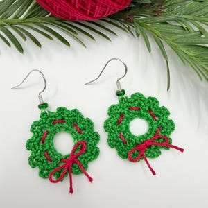 Può includere: Un paio di orecchini a forma di ghirlanda all'uncinetto verde fatti a mano con dettagli e fiocchi rossi. Gli orecchini hanno ganci color argento e piccole perline verdi. Un gomitolo di filo rosso e ramoscelli verdi sullo sfondo.
