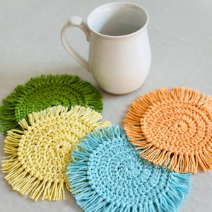May include: Four crocheted coasters with fringe in green, yellow, blue, and orange. The coasters are arranged in a circle around a white ceramic mug.