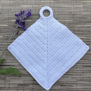 Pode incluir: Um pegador de panela de crochê branco com uma alça de laço. O pegador de panela está sobre um fundo marrom tecido.