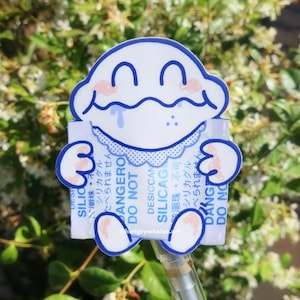 Peut inclure: Un autocollant blanc et bleu représentant un personnage de dessin animé tenant un rectangle avec du texte. Le personnage a un visage souriant avec des accents roses. Le rectangle indique "DESICCANT SILICAGEL DO NOT" avec du texte japonais.