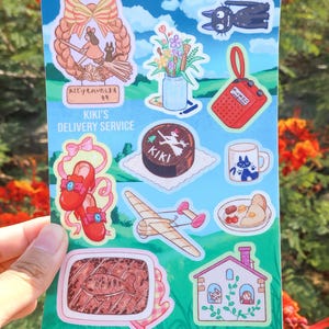 May include: A sticker sheet with illustrations from Kiki's Delivery Service. The sheet includes images of Kiki on a broom, a cake, a radio, a mug, a plane, a house, and other themed elements. The background is blue.