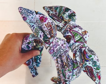 May include: A patterned fabric headband with a bow. The headband displays a print with various illustrations and text in multiple colours, including pink, green, and blue. The headband is held by a hand against a white background.
