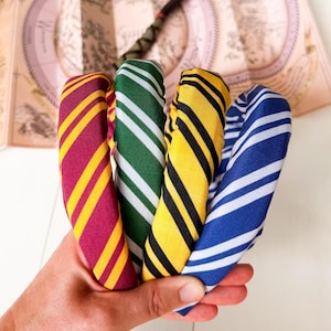 May include: Four fabric headbands featuring diagonal stripes in maroon and gold, green and silver, yellow and black, and blue and silver. A wooden wand and a folded parchment map with intricate illustrations are positioned in the background.