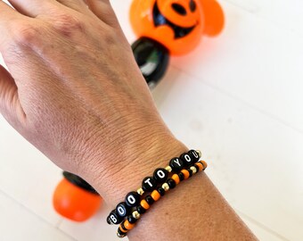 May include: Two Halloween-themed bracelets with the words "BOO TO YOU" spelled out in black and white letter beads. The bracelets feature orange and gold beads. An orange Mickey Mouse-shaped Halloween decoration is in the background.