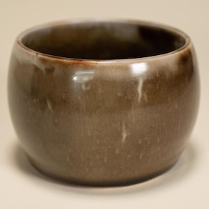 May include: A handcrafted, round ceramic bowl with a glossy, brown glaze. The bowl has a smooth, curved shape and a slightly lighter rim. The interior is dark brown, and the exterior has subtle variations in color, creating a unique, earthy aesthetic.