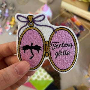 Peut inclure: Écusson brodé avec un design ovale rose, représentant une silhouette de dragon noir et les mots "Fantasy girlie" en écriture cursive. L'écusson a une bordure dorée et un nœud violet en haut.