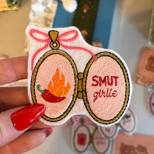 Könnte beinhalten: Gestickter Aufnäher mit rosa Hintergrund und goldenem Rand. Eine Seite zeigt eine rote Chilischote mit Flammen, die andere Seite liest "SMUT girlie" in Rot. Eine rosa Schleife und ein Faden verbinden die beiden Seiten.