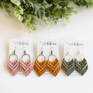 May include: Three pairs of macrame earrings in pink, mustard yellow, and olive green. Each pair features a woven design with a circular hoop and fringe. The earrings are displayed on white cards with the text "Mended Fibers".