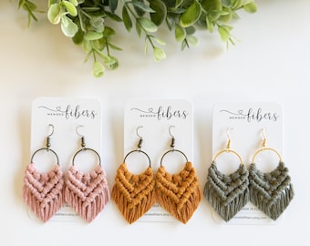 May include: Three pairs of macrame earrings in pink, mustard yellow, and olive green. Each pair features a woven design with a circular hoop and fringe. The earrings are displayed on white cards with the text "Mended Fibers".