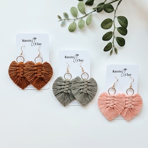May include: Three pairs of heart-shaped tassel earrings in rust, sage green, and blush pink. Each pair is made with a gold hoop and a knotted tassel. The earrings are displayed on white cards with the text "Knots & Clay".