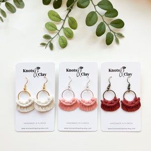 May include: Three pairs of handmade macrame earrings with different colored tassels. The earrings are displayed on white cards with the text "Knots & Clay" and "Handmade in Florida" along with the website address.