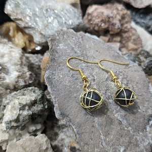 May include: A pair of gold wire wrapped earrings with black stones. The earrings are hanging from a rock background.