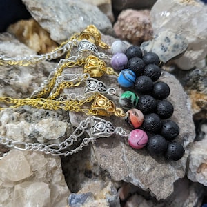 Op de afbeelding: Een set van zeven kettingen met zilveren en gouden schakels en lavastenen kralen. Elke ketting heeft een anders gekleurde glazen kraal met een swirl-patroon. De kettingen worden getoond op een bed van stenen.