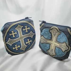 May include: Two blue coin purses with a decorative cross design. The purses have a textured appearance and a zipper closure. The cross is in a lighter color, with a detailed, ornate design. The background is a light blue color.