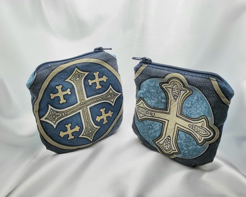 May include: Two blue coin purses with a decorative cross design. The purses have a textured appearance and a zipper closure. The cross is in a lighter color, with a detailed, ornate design. The background is a light blue color.
