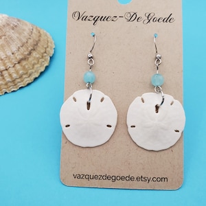 Op de afbeelding: Witte zanddollar oorbellen met een lichtblauwe kraal en zilverkleurige haken. De oorbellen worden gepresenteerd op een bruine kaart met de tekst "Vazquez-De Goede" en "vazquezdegoede.etsy.com" op een blauwe achtergrond.