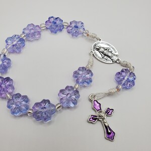 Könnte beinhalten: Ein Rosenkranz mit durchscheinenden, blütenförmigen Perlen in Lila- und Blautönen. Der Rosenkranz hat ein silberfarbenes Kruzifix mit lila Emaille und einen silbernen ovalen Anhänger mit einer religiösen Figur. Die Perlen sind auf einer weißen Schnur aufgefädelt.