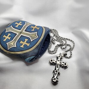May include: A silver cross pendant with an ornate design hangs from a silver chain. A blue pouch with a cross design is next to the necklace. The pouch has a zipper and a gold border.