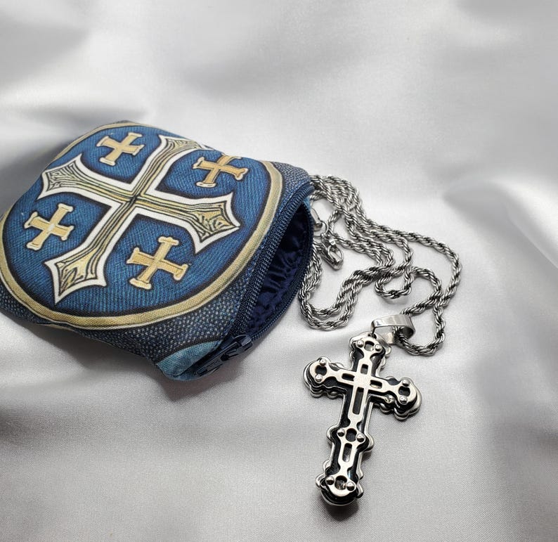 May include: A silver cross pendant with an ornate design hangs from a silver chain. A blue pouch with a cross design is next to the necklace. The pouch has a zipper and a gold border.