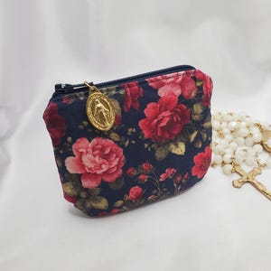 May include: A small fabric pouch featuring a dark navy blue background with a red rose floral pattern. The pouch has a black zipper closure with a gold-toned oval medal charm attached to the zipper pull. A white beaded rosary is visible nearby.