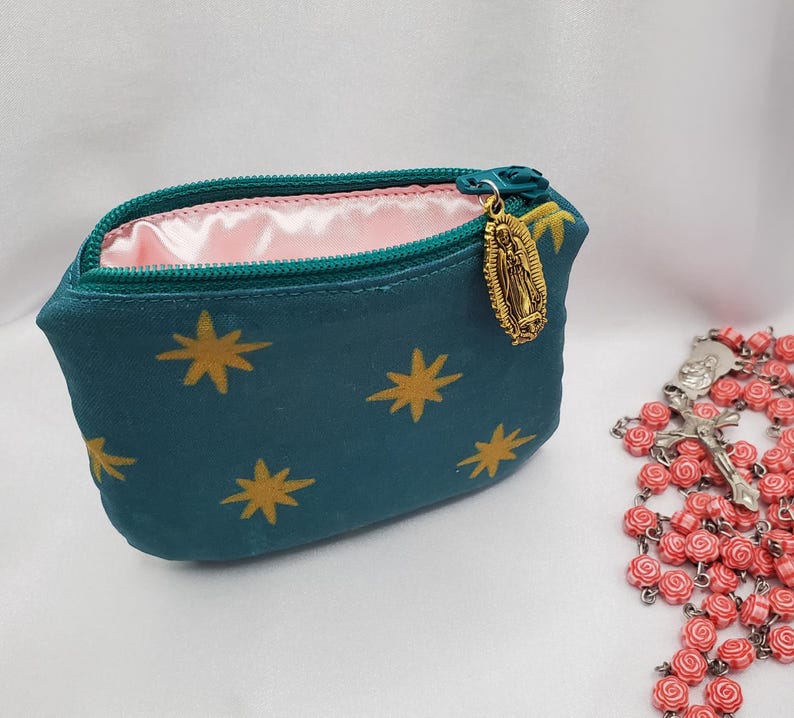 May include: Teal zippered pouch with gold starburst pattern and a gold Virgin Mary charm. The interior is pink. A rosary with pink rose-shaped beads is visible on the right.