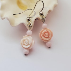 May include: A pair of dangle earrings featuring carved light pink and cream rose-shaped beads. Each earring is suspended from a silver-toned metal hook and includes small, round, pale pink accent beads above and below the floral centerpiece.