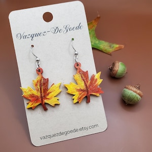 May include: Hand-painted maple leaf earrings in autumn colors of yellow, orange, and red. The earrings hang from silver-colored hooks. The earrings are displayed on a tan card with the text "Vazquez-De Goede" and "vazquezdegoede.etsy.com".