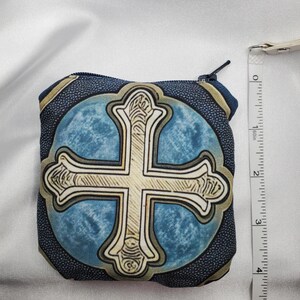 May include: A small, square coin purse with a cross design. The purse features a blue background with a gold cross in the center. The purse is approximately 4 inches tall and has a zipper closure. The edges of the purse are dark blue with a gold trim.
