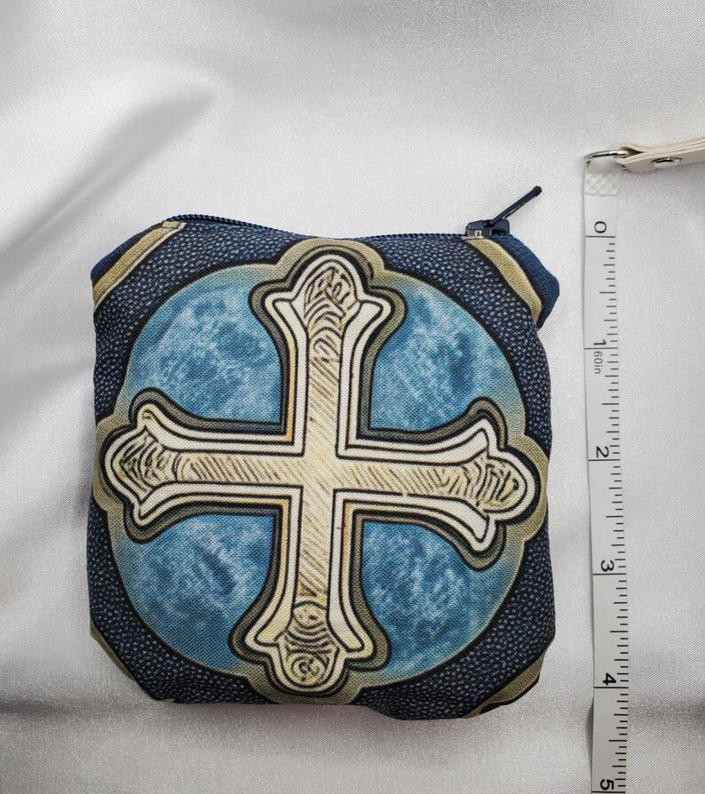 May include: A small, square coin purse with a cross design. The purse features a blue background with a gold cross in the center. The purse is approximately 4 inches tall and has a zipper closure. The edges of the purse are dark blue with a gold trim.