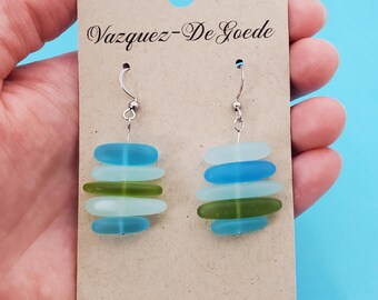 May include: Handmade earrings featuring stacks of sea glass in shades of blue and green, suspended from silver-tone hooks. The earrings are displayed on a brown card with the text "Vazquez-De Goede" and "vazquezdegoede.etsy.com".