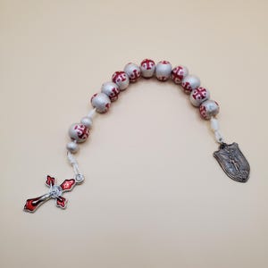 May include: A rosary with silver-colored beads, each adorned with a red cross design. The rosary features a red and silver cross pendant and a shield-shaped medal with an engraved figure.