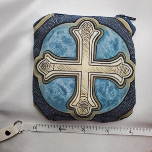 May include: A small, square coin purse with a blue and beige cross design. The cross is centered on a light blue background, framed by a dark blue border. The purse has a zipper closure and measures approximately 4 inches wide.