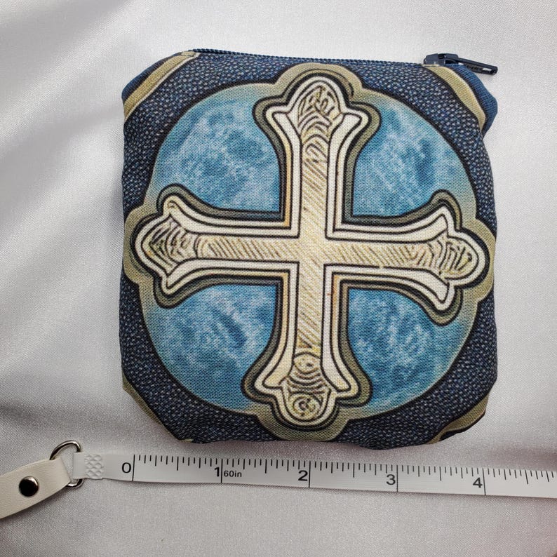 May include: A small, square coin purse with a blue and beige cross design. The cross is centered on a light blue background, framed by a dark blue border. The purse has a zipper closure and measures approximately 4 inches wide.