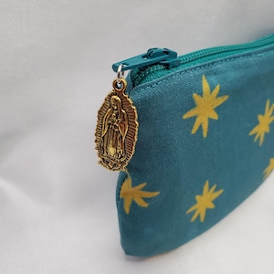May include: Teal pencil case with a gold zipper and a gold Virgin Mary charm. The case has a pattern of gold starbursts. The zipper is teal, matching the case. The charm is oval-shaped and detailed.