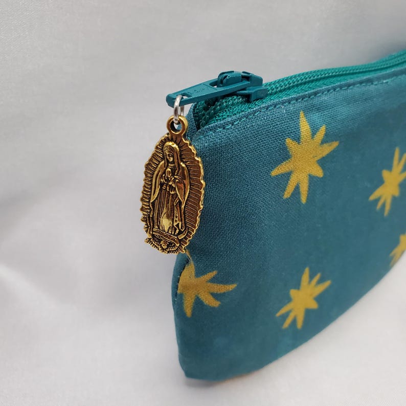 May include: Teal pencil case with a gold zipper and a gold Virgin Mary charm. The case has a pattern of gold starbursts. The zipper is teal, matching the case. The charm is oval-shaped and detailed.