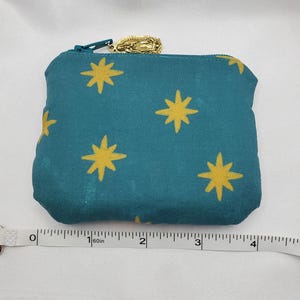 May include: Teal fabric zippered pouch with a gold star pattern. The pouch has a gold zipper pull with a religious charm. The pouch is approximately 4 inches wide.