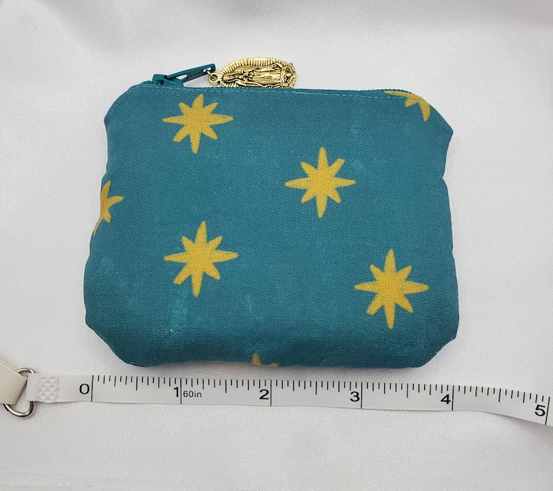 May include: Teal fabric zippered pouch with a gold star pattern. The pouch has a gold zipper pull with a religious charm. The pouch is approximately 4 inches wide.