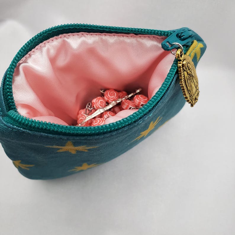 May include: Teal zippered pouch with gold star pattern and a pink satin lining. The pouch contains a rosary with red and white beads and a silver crucifix. A gold-colored charm hangs from the zipper.