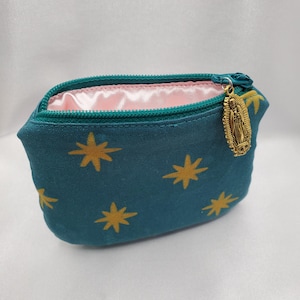 May include: Teal cosmetic bag with a gold star pattern and a gold zipper pull featuring a religious figure. The interior is lined with pink fabric. The bag is designed to hold makeup or other small items.