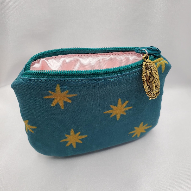 May include: Teal cosmetic bag with a gold star pattern and a gold zipper pull featuring a religious figure. The interior is lined with pink fabric. The bag is designed to hold makeup or other small items.
