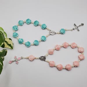 May include: Two rosaries with shell-shaped beads. One is light blue, the other is pink. Each has a silver cross and a center piece. The beads are strung on a cream-colored cord. The background is white.