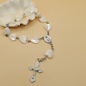 May include: A rosary with heart-shaped beads and a silver crucifix. The beads are white and pearlescent, strung together with silver-colored links. The crucifix and center medal feature religious imagery. The rosary is displayed on a beige surface next to a seashell.