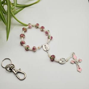 May include: A rosary featuring pink and white floral beads, a silver Virgin Mary pendant, and a pink cross pendant. A silver keychain is also present. The rosary is made of white cord.