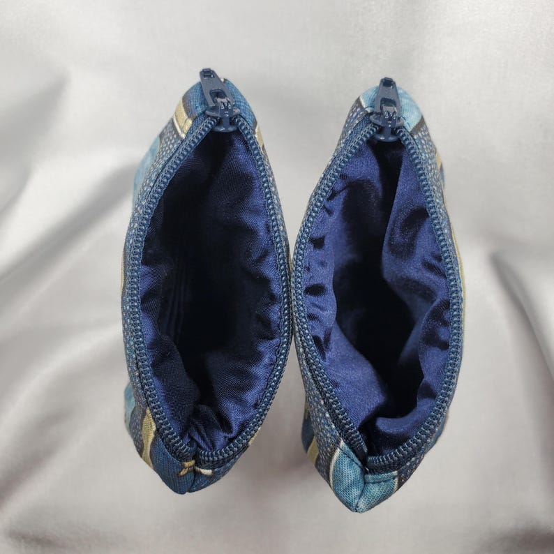 May include: Two small, open zippered pouches. The exterior fabric is a blue and white pattern with gold stripes. The interior is a solid navy blue satin. The zippers are blue.