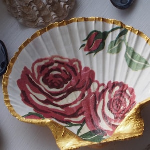 May include: A decorative scallop shell dish featuring a painted design of two large red roses and a single rosebud with green leaves. The interior of the shell is white, while the scalloped edges and the base are painted with a metallic gold finish.