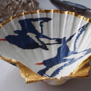 May include: A large, white scallop shell bowl featuring a hand-painted design of two dark blue birds in flight with small orange beaks. The scalloped edges of the shell are finished with a metallic gold trim, highlighting the natural ridges.