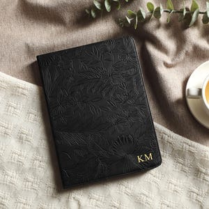 Könnte beinhalten: Ein schwarzes Lederjournal mit geprägtem floralem Design und goldenen Initialen "KM" wird gezeigt. Eine Tasse Tee auf einer weißen Untertasse befindet sich im Hintergrund, mit grünem Laub. Das Journal liegt auf einer cremefarbenen Oberfläche.